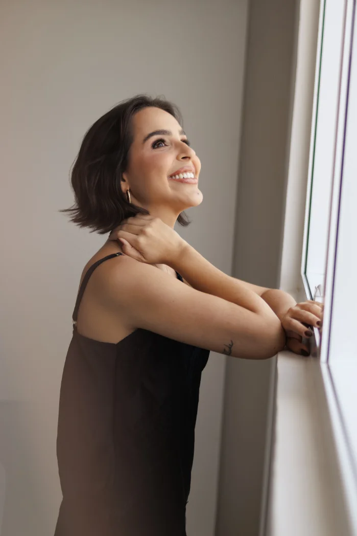 Dra. Duda Carreira sorrindo, apoiada no parapeito de uma janela